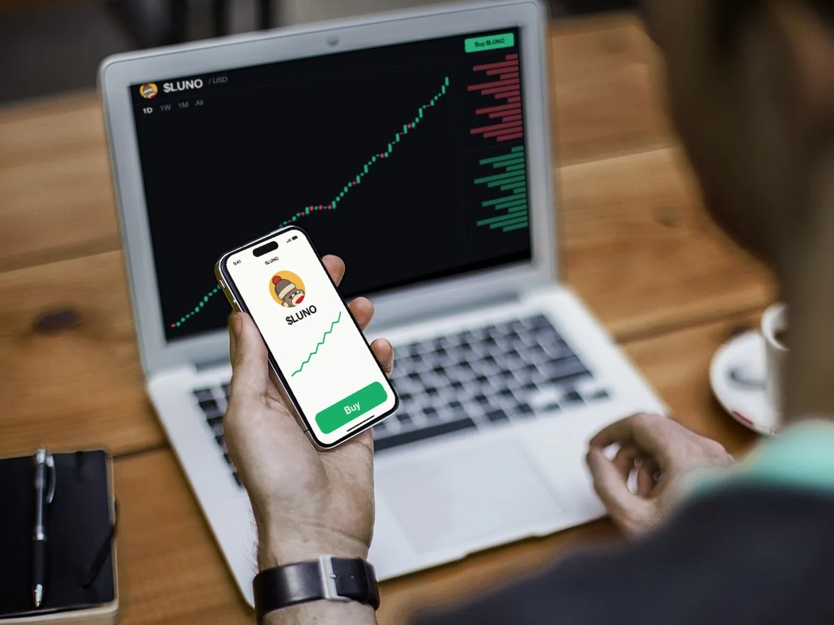 A laptop showing the $LUNO chart climbing, and an iPhone in his hand open on a Buy button